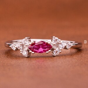 Op de afbeelding: Een zilveren ring met een marquise-vormige robijn, omringd door kleinere witte edelstenen. De ring ligt op een bruin oppervlak.