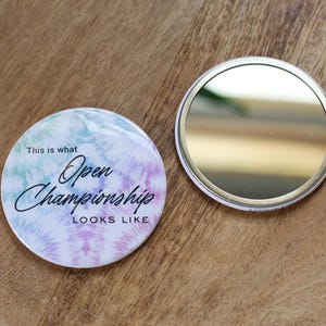 May include: Two round, tie-dye style pinback buttons on a wooden surface. One button has a mirror finish, the other has the text "This is what Open Championship LOOKS LIKE" in black script over a pastel background.