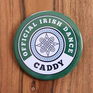 May include: A round, green and white button with the text "OFFICIAL IRISH DANCE" and "CADDY" in white. The center features a Celtic knot design in white and blue, surrounded by a laurel wreath.