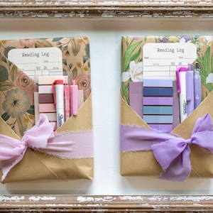 Two wrapped book packages in kraft and floral paper displayed in a rustic wooden tray, each styled with soft purple or pink ribbon and coordinated pens, sticky tabs, and reading logs.