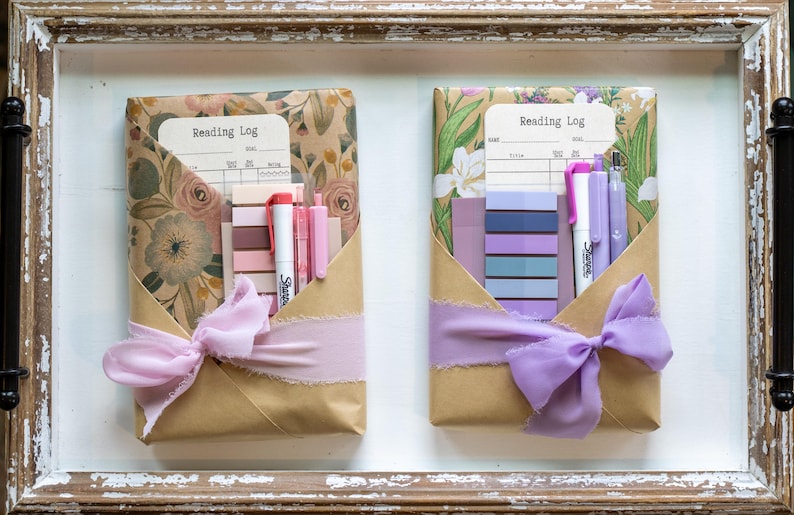Two wrapped book packages in kraft and floral paper displayed in a rustic wooden tray, each styled with soft purple or pink ribbon and coordinated pens, sticky tabs, and reading logs.