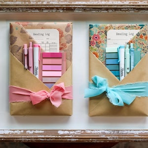 Two vibrant blind date book packages with pink and turquoise ribbons and matching sticky tabs, wrapped in floral kraft paper, and displayed in a vintage-style wood tray.