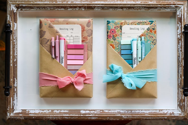 Two vibrant blind date book packages with pink and turquoise ribbons and matching sticky tabs, wrapped in floral kraft paper, and displayed in a vintage-style wood tray.