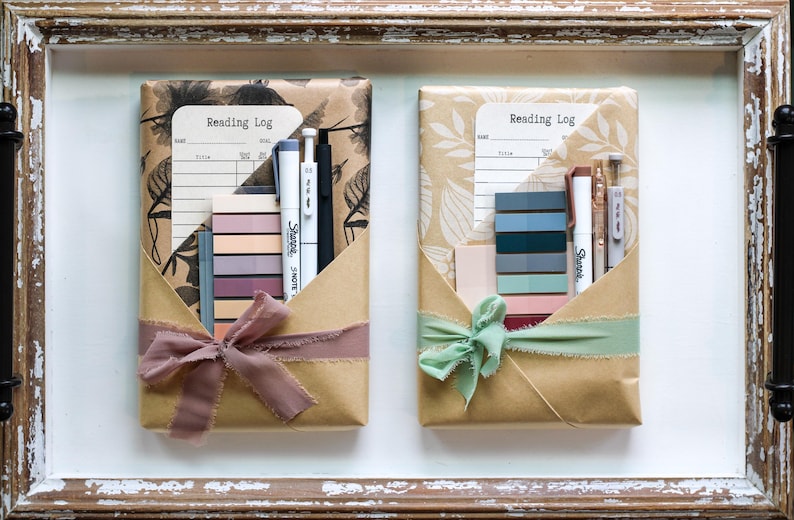 Two kraft-wrapped book kits with dark floral and leaf designs, each with moody-toned stationery, pens, sticky notes, and reading logs—presented inside a distressed wooden tray.
