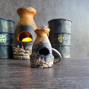 Peut inclure: Un porte-clés en forme de four miniature en argile avec une base en pierre texturée, un corps arrondi beige avec des marques noires semblables à de la suie et une petite ouverture circulaire. Il comporte un anneau en métal argenté.