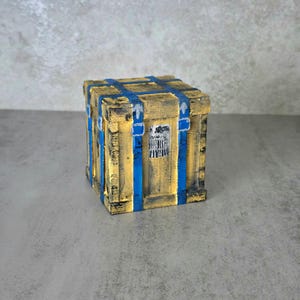 May include: A square wooden crate with a weathered yellow finish and black distressed detailing. The box features prominent blue painted straps running vertically and horizontally across its sides, with a small silver rectangular label on the front.