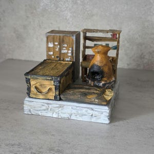 May include: A miniature diorama featuring a wooden cabinet, a chest with a curved handle, a rustic open shelf unit, and a central orange clay oven with a dark opening. The entire assembly is mounted on a textured white stone-like base.