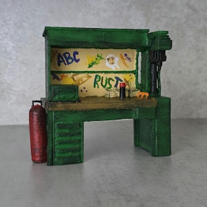 May include: A miniature green workbench featuring a textured finish, a brown wooden-look surface, and a red cylindrical tank attached to the side. The back panel displays painted text reading ABC and RUST alongside colorful abstract shapes.