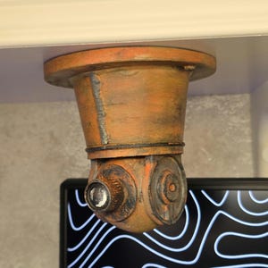 Può includere: Un supporto da soffitto in stile industriale color arancione e marrone invecchiato, con base cilindrica e alloggiamento rotante simile a una telecamera. L'oggetto presenta una finitura testurizzata e usurata con accenti scuri e una lente.