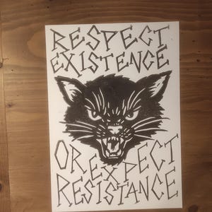 Op de afbeelding: Zwart-wit illustratie op papier met een woest kattengezicht met de tekst "Respect Existence Or Expect Resistance". De kat heeft een boze uitdrukking met ontblote tanden. Het kunstwerk staat op een houten oppervlak.