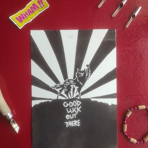 Puede incluir: Ilustración en blanco y negro de un lobo aullando contra un diseño de rayos de sol, con el texto "GOOD LUCK OUT THERE". La imagen está sobre una superficie roja, con una pegatina "WHAM!!", pequeñas herramientas plateadas y una pulsera de cuentas.