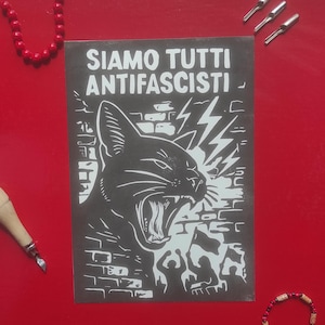 Può includere: Una stampa in bianco e nero con la scritta "SIAMO TUTTI ANTIFASCISTI" e un'illustrazione di un gatto che ruggisce. La stampa è su una superficie rossa, con perline rosse e altri piccoli oggetti intorno.