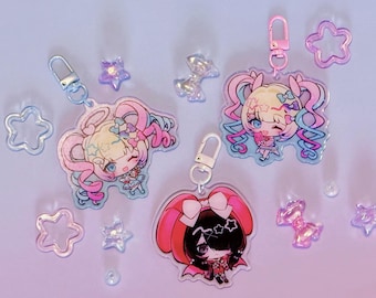 Puede incluir: Tres llaveros de acrílico con personajes de estilo anime con cabello y atuendos coloridos, cada uno unido a un cierre de metal. El conjunto incluye pequeñas estrellas y lazos decorativos de plástico en tonos rosa, azul y transparente.
