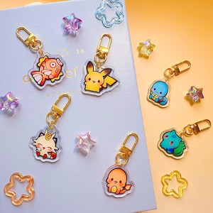 Puede incluir: Una colección de llaveros acrílicos coloridos con personajes de dibujos animados, incluyendo un pez y Pikachu. Cada llavero tiene un cierre dorado. Elementos decorativos en forma de estrella y flor están dispersos.