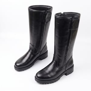 May include: A pair of black knee-high boots featuring a smooth finish, a rounded toe, a side zipper closure, and a rectangular hook-and-loop strap at the top. The boots have a thick, lugged rubber sole and a low block heel for traction.