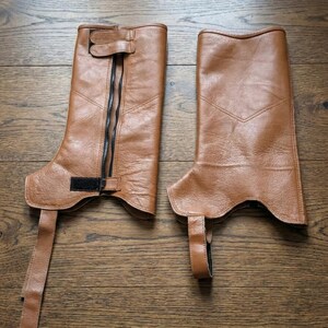Puede incluir: Un par de chaps de cuero marr&oacute;n, con cierre de cremallera y detalles de hebilla. Los chaps tienen un dise&ntilde;o hasta la rodilla con una correa y hebilla en la parte superior e inferior. El cuero tiene un aspecto ligeramente desgastado.