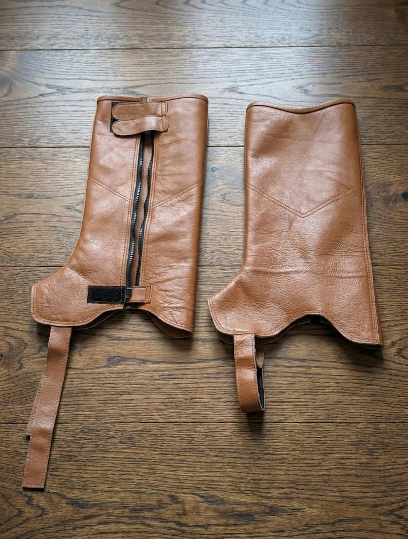 Puede incluir: Un par de chaps de cuero marr&oacute;n, con cierre de cremallera y detalles de hebilla. Los chaps tienen un dise&ntilde;o hasta la rodilla con una correa y hebilla en la parte superior e inferior. El cuero tiene un aspecto ligeramente desgastado.