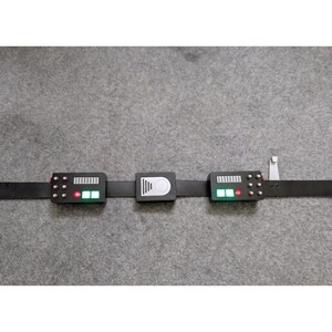 May include: A black utility belt featuring three rectangular modules attached to the strap. The two outer modules have small silver buttons, a silver grid, and illuminated green and red lights. The center module has a silver circular design.