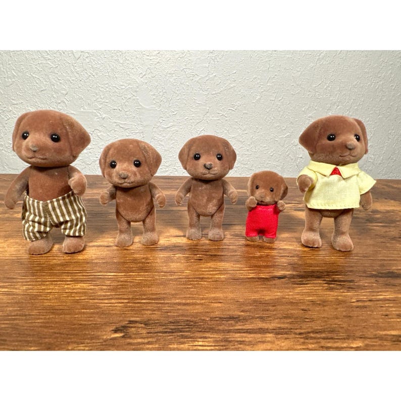 Op de afbeelding: Vijf bruine speelgoedhondfiguren. E&eacute;n hond draagt een gestreepte broek, een andere een geel shirt met een rode kraag en een rode overall. De andere twee honden staan rechtop.