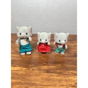 May include: Three gray elephant figurines wearing different outfits. One is in teal shorts, another in a red dress, and the third in green and white checkered overalls. The toys are on a wooden surface.
