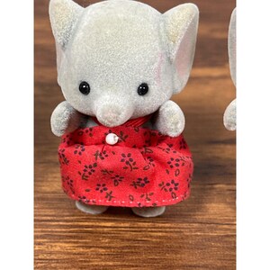 May include: A gray elephant figurine wearing a red dress with a floral pattern. The figurine has black eyes and a small trunk. The dress is buttoned at the neck. The toy is part of a collection of animal figurines.