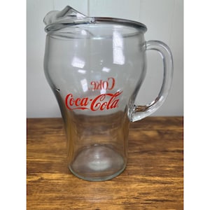 Peut inclure: Pichet Coca-Cola en verre transparent avec une anse et un couvercle. Le logo Coca-Cola est imprimé en rouge sur le devant du pichet. Le pichet est posé sur une surface en bois.