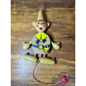 May include: A vintage wooden pull-string toy in the shape of a whimsical figure. The figure has a painted face, a pointed hat, and a green and yellow outfit. The arms, legs, and head are articulated, and the toy is painted in bright colors.