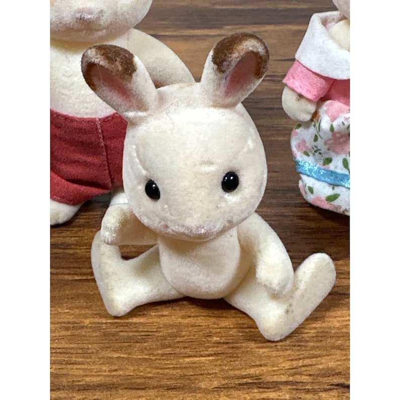 Pu&ograve; includere: Una statuetta di coniglio bianco con orecchie con la punta marrone e occhi neri &egrave; seduta con le gambe divaricate. Il coniglio fa parte di una collezione di piccole statuette di animali, con altre figure parzialmente visibili sullo sfondo.