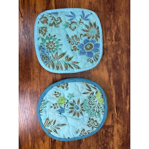 May include: Two quilted pot holders with a floral pattern. One is square with rounded corners, and the other is round. Both have a light blue background with brown, blue, and green flowers and a blue border.