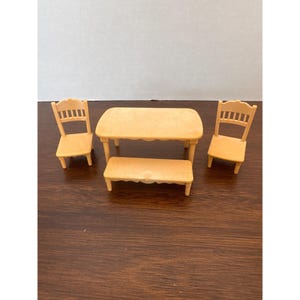 May include: A miniature dollhouse dining set in a light tan color. The set includes a table, a bench, and two chairs. The table has a scalloped edge, and the chairs have a ladder-back design. The bench is rectangular.