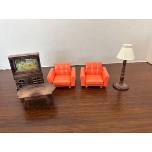 Puede incluir: Juego de muebles en miniatura para casa de muñecas con dos sillones naranjas, un mueble de TV de madera, una mesa pequeña y una lámpara de pie con una pantalla crema. La pantalla del televisor muestra una escena con animales. El conjunto está dispuesto sobre una superficie de madera.