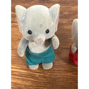 May include: A gray elephant figurine wearing teal shorts. The elephant has large ears, black eyes, and a trunk. The figurine is made of a soft, flocked material. A second elephant figurine is partially visible.