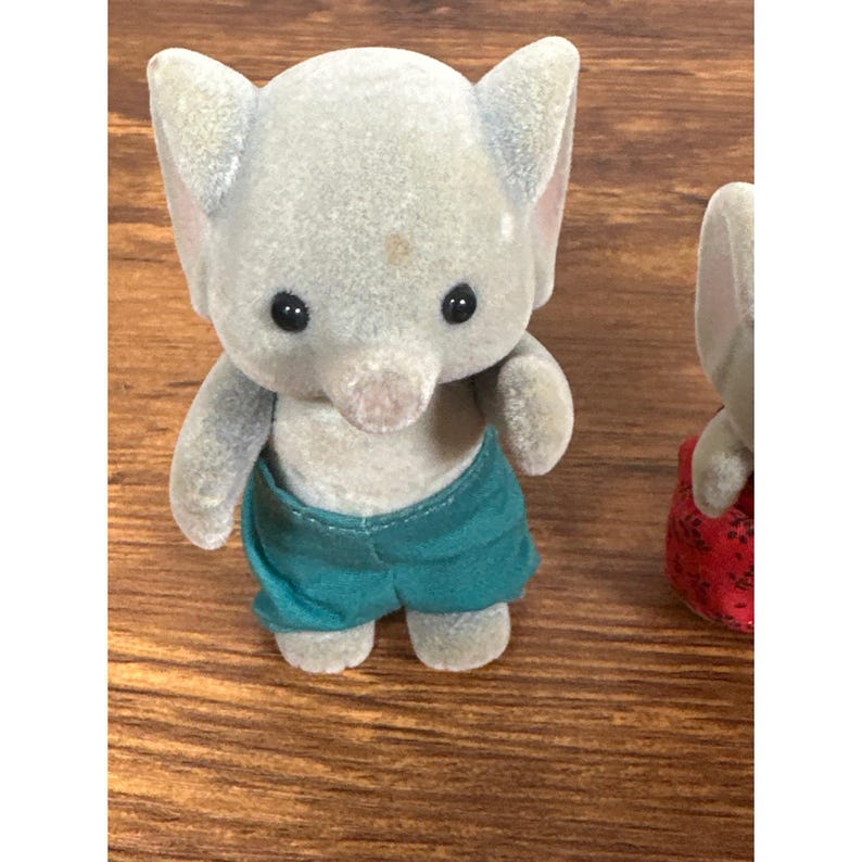 May include: A gray elephant figurine wearing teal shorts. The elephant has large ears, black eyes, and a trunk. The figurine is made of a soft, flocked material. A second elephant figurine is partially visible.