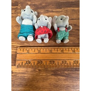 May include: Three gray elephant figurines wearing teal shorts, a red dress, and green checkered overalls. A wooden ruler with inch markings is in the foreground. The figurines are standing on a wooden surface.