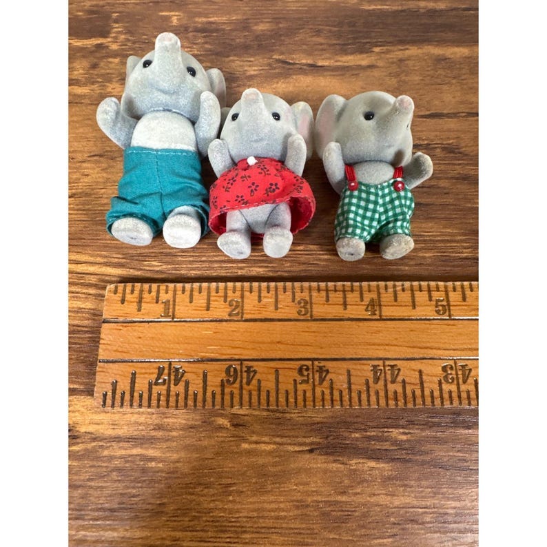 May include: Three gray elephant figurines wearing teal shorts, a red dress, and green checkered overalls. A wooden ruler with inch markings is in the foreground. The figurines are standing on a wooden surface.