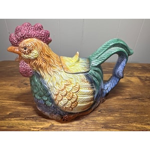 Könnte beinhalten: Eine keramische Teekanne in Form eines Hahns mit einem lebendigen Farbschema. Die Teekanne hat einen roten Kamm, orange und gelbe Federn sowie einen grünen Schwanz und Griff. Die Teekanne steht auf einer Holzoberfläche.