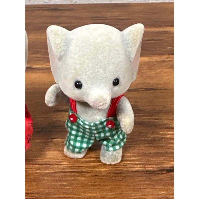 May include: A gray elephant figurine wearing green and white checkered overalls with red suspenders. The figurine has black eyes and a pink trunk. The toy is a collectible item.