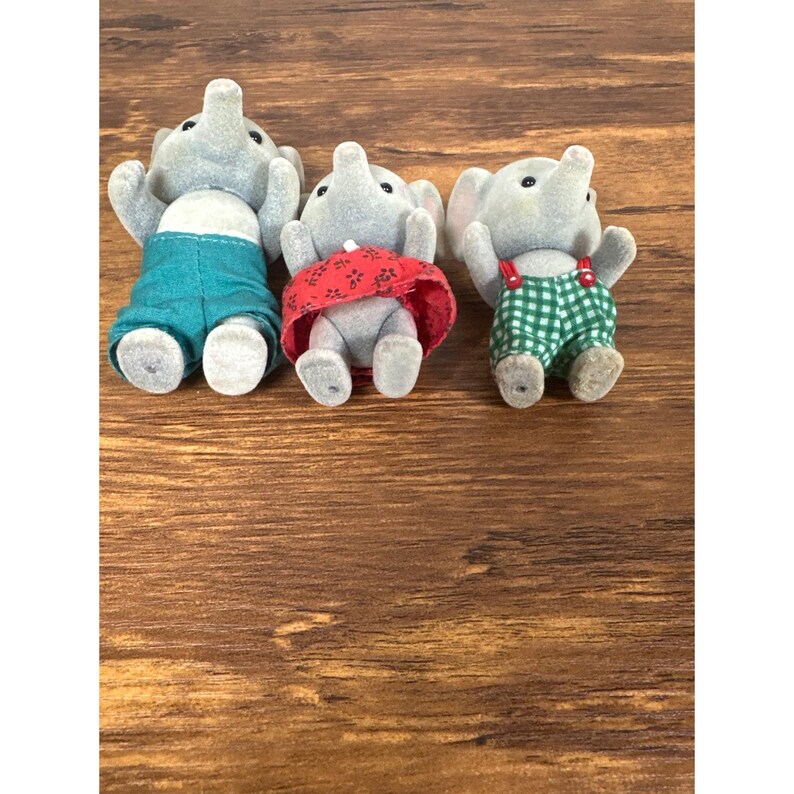 May include: Three gray elephant figurines wearing different outfits. One elephant is wearing teal shorts, another a red skirt, and the third has green and white checkered shorts with red suspenders. The figurines are on a wooden surface.
