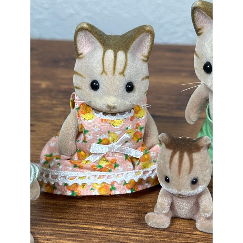 Może przedstawiać: Grupa figurek kot&oacute;w Calico Critters. Jeden kot ma na sobie r&oacute;żową sukienkę z białym koronkowym wykończeniem i wzorem ż&oacute;łtych i pomarańczowych owoc&oacute;w. Pozostałe koty są w r&oacute;żnych pozach i strojach. Figurki mają brązowe i jasnobrązowe futro.