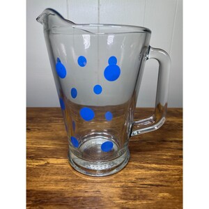 Puede incluir: Jarra de vidrio transparente con asa y boquilla, decorada con lunares azules. La jarra está diseñada con una base ancha y una parte superior más estrecha, adecuada para servir bebidas. La jarra es de vidrio transparente.