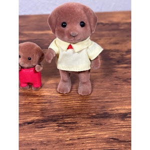 Op de afbeelding: Twee bruine speelgoedhondenfiguren. E&eacute;n hond draagt een lichtgeel shirt met een rode kraag. De andere hond draagt een rode overall. Het speelgoed staat op een houten oppervlak.