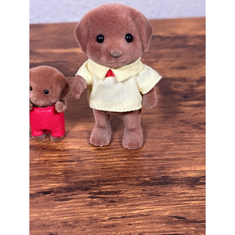 Op de afbeelding: Twee bruine speelgoedhondenfiguren. E&eacute;n hond draagt een lichtgeel shirt met een rode kraag. De andere hond draagt een rode overall. Het speelgoed staat op een houten oppervlak.
