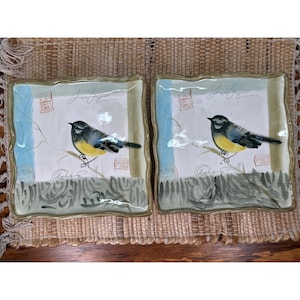 May include: Two square ceramic plates with a bird design. Each plate features a blue, yellow, and black bird perched on a branch against a white background with blue and green accents. The plates have a wavy edge and a gold trim.
