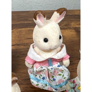 Pu&ograve; includere: Una piccola statuetta di coniglio color crema con orecchie con punte marroni e occhi neri. Il coniglio indossa un vestito rosa e bianco con un motivo floreale e dettagli di nastro blu. La statuetta fa parte di una collezione.