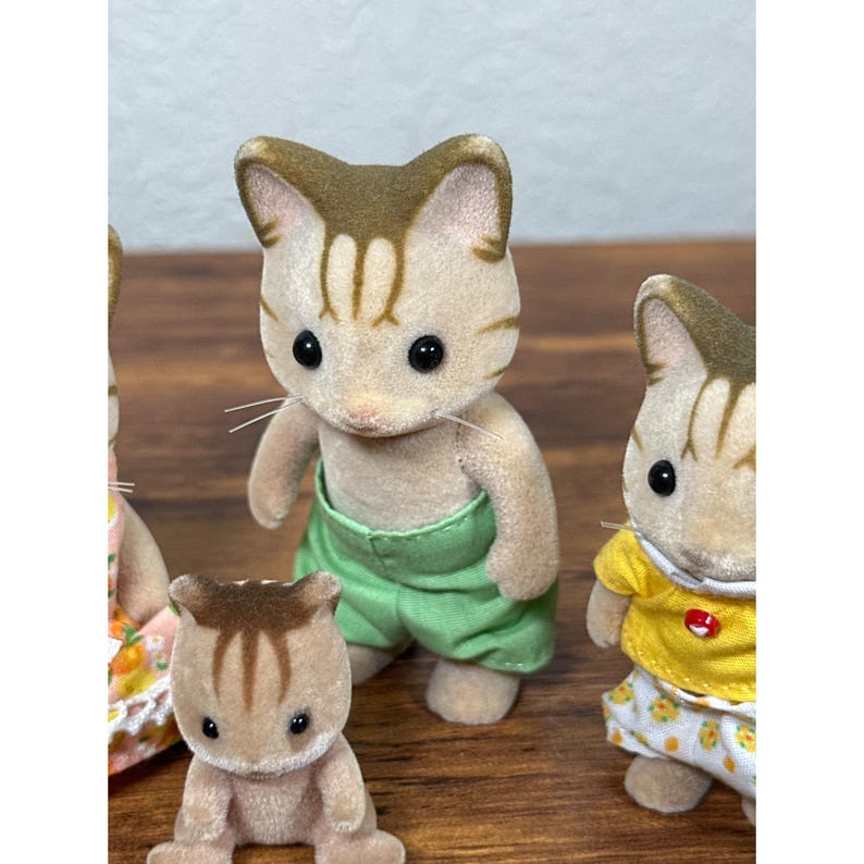 Może przedstawiać: Grupa miniaturowych figurek kot&oacute;w. Centralny kot ma na sobie zielone szorty. Koty mają brązowe i beżowe futro z czarnymi oczami. Jeden kot ma na sobie ż&oacute;łtą koszulę i kwiecistą sp&oacute;dnicę. Figurki mają miękką, flokowaną teksturę.