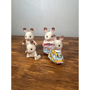 Pu&ograve; includere: Quattro piccole statuette di conigli color crema con orecchie con la punta marrone. Due conigli sono in piedi, uno &egrave; seduto e uno &egrave; in posizione di gattonamento. Due conigli indossano abiti con motivi floreali. Le statuette sono su una superficie di legno.