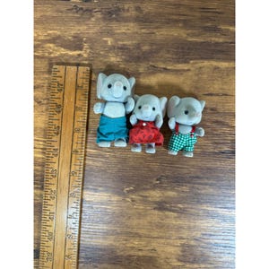 May include: Three gray elephant figurines wearing colorful outfits. One elephant is in teal pants, another in a red dress with white polka dots, and the third in green and white checkered overalls. A wooden ruler is on the left.