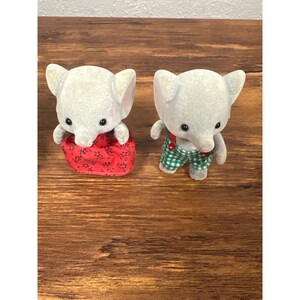 May include: Two gray elephant figurines. One wears a red floral print dress, the other wears green and white checkered overalls with red suspenders. Both have black eyes and large ears. The figurines are on a brown wooden surface.