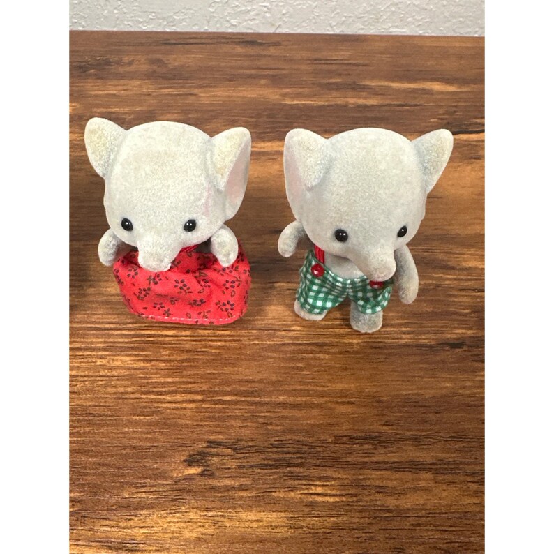 May include: Two gray elephant figurines. One wears a red floral print dress, the other wears green and white checkered overalls with red suspenders. Both have black eyes and large ears. The figurines are on a brown wooden surface.