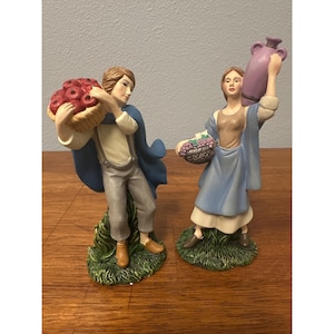 May include: Two ceramic figurines of figures in period dress. One carries a basket of red apples, the other a basket of purple grapes and a purple jug. Both stand on green, grassy bases.